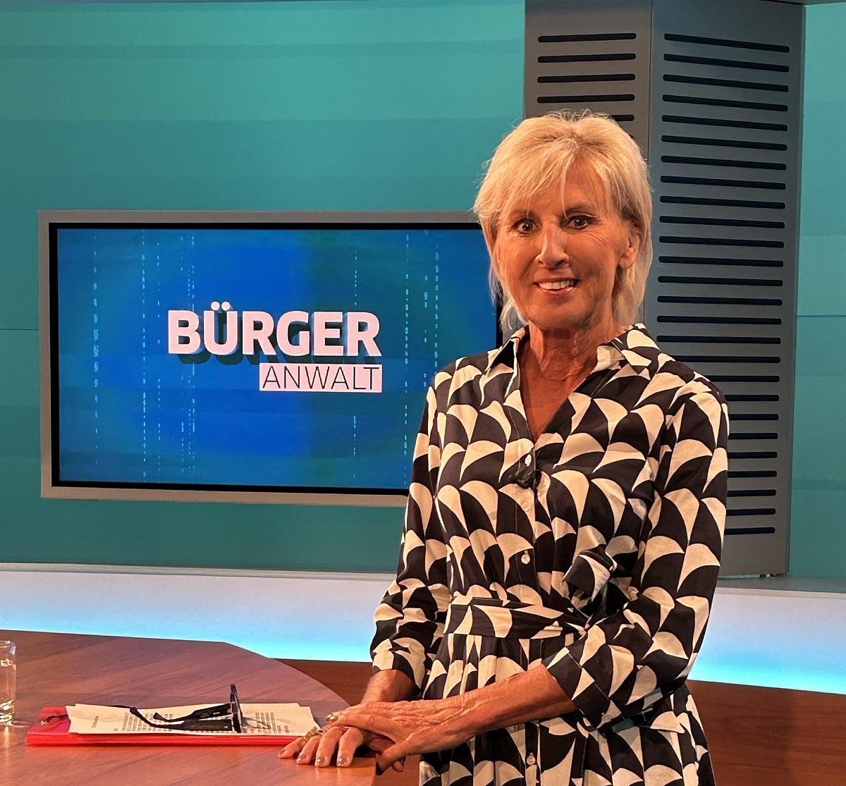 Volksanwältin Gaby Schwarz in einem schwarz-weißen Kleid, stehend an einem Tisch im TV Studio.