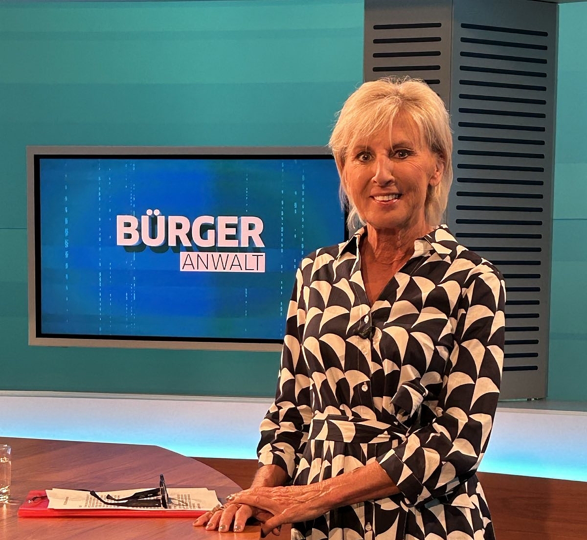 People's Advocate Gaby Schwarz in a black and white dress, standing at a table in the TV studio.