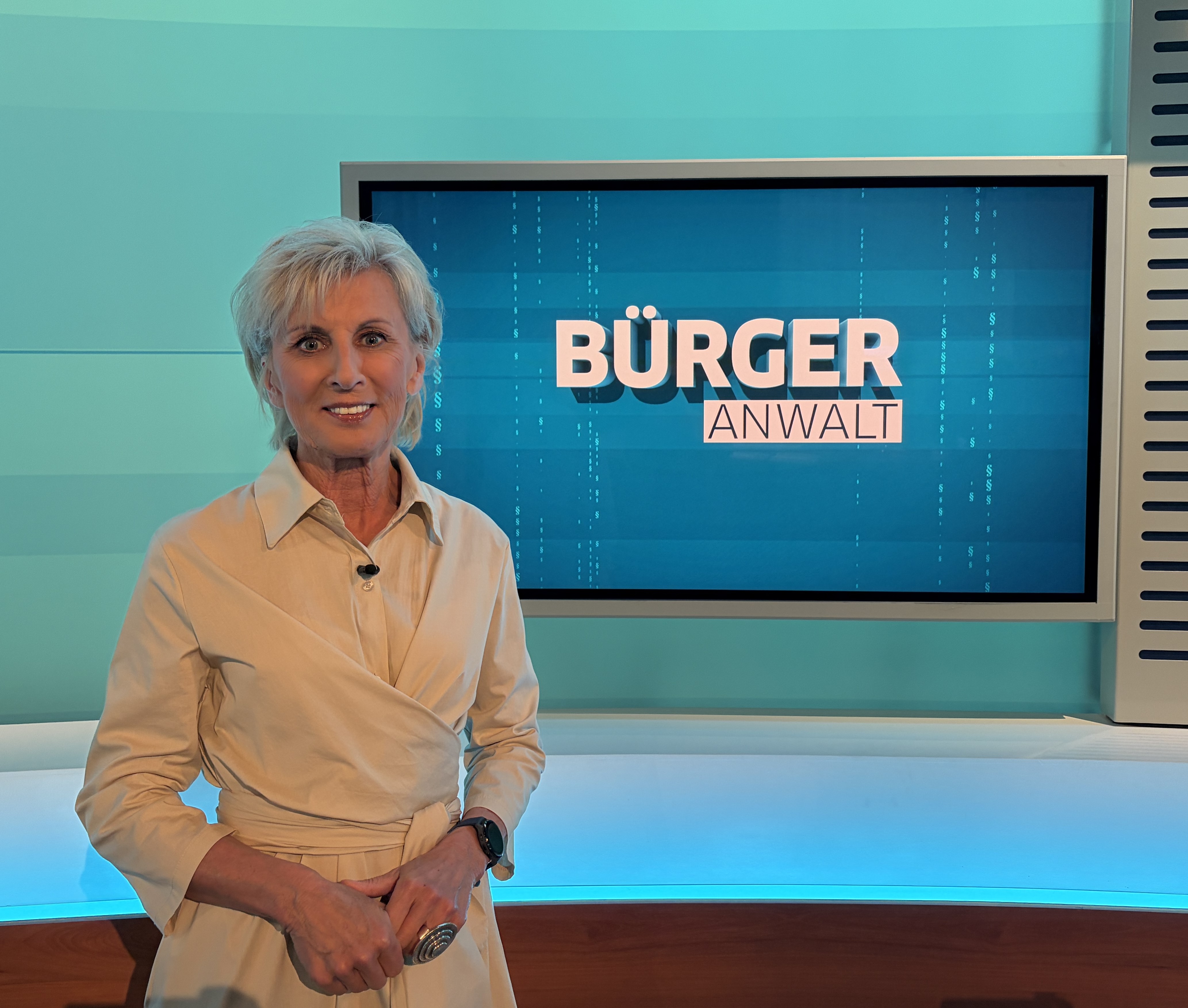Volksanwältin Gaby Schwarz steht in einem beigen Kleid im ORF Studio vor dem Bürgeranwalt Logo.