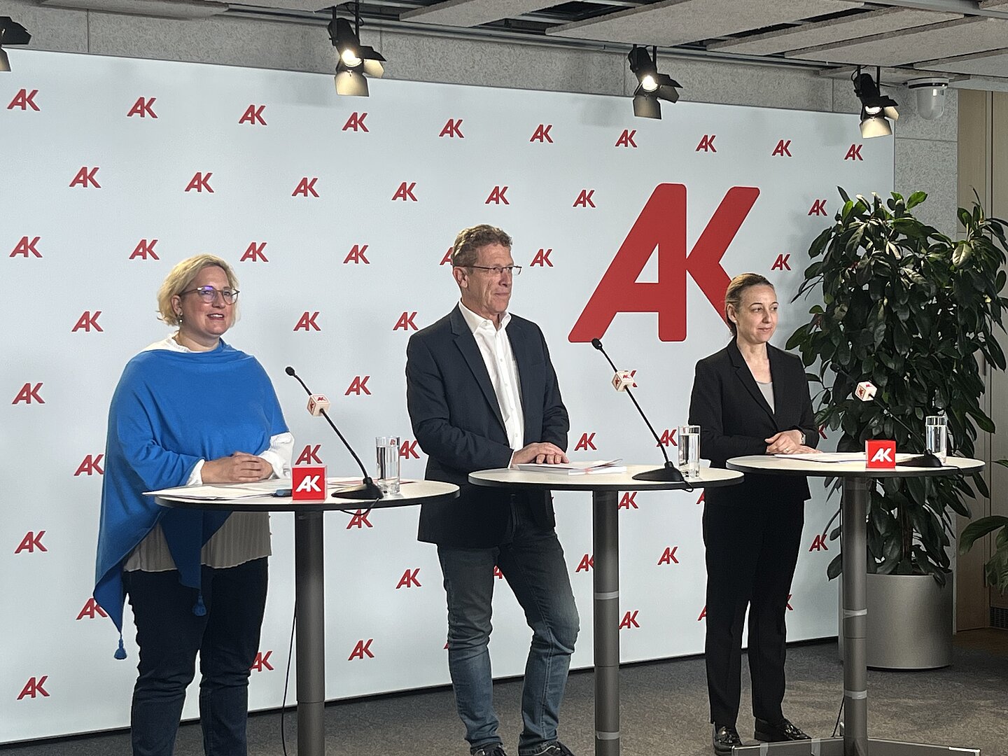 Zwei Frauen und ein Mann stehend am Podium mit Mikrophonen vor ihnen auf Stehtischen im Hintergrund die Pressewand der Arbeiterkammer.