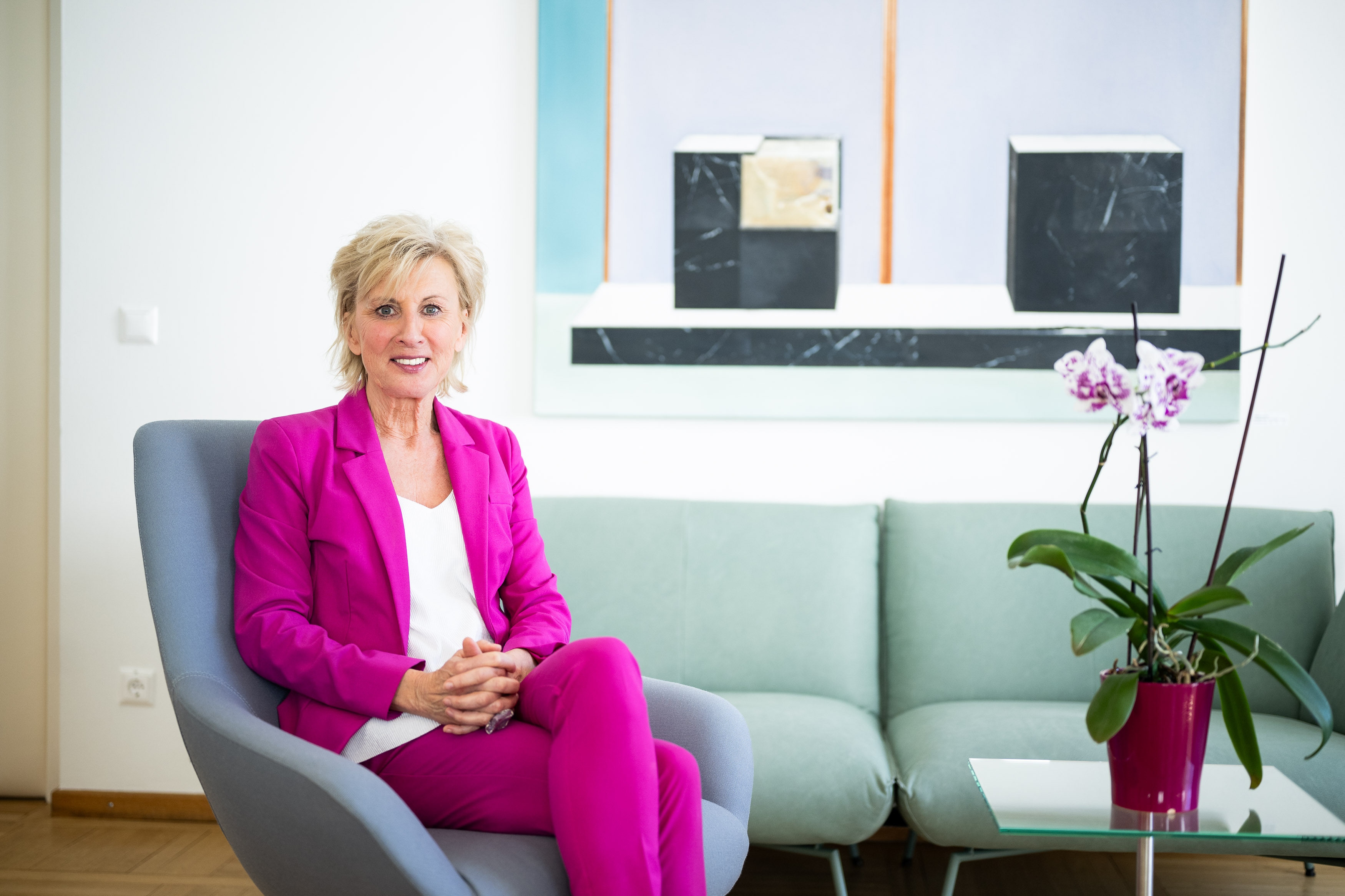 Volksanwältin Gaby Schwarz trägt einen pinken Hosenanzug und sitzt in einem türkisen Fauteuil