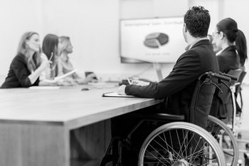 Besprechung im Büro, sieben Personen sitzend bei Konferenztisch, teilweise unscharf, ein Mann davon im Rollstuhl, Präsentation im Hintergrund mit Kreisdiagramm.