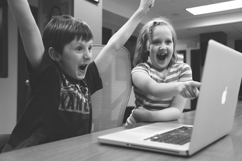Zwei Kinder vor Laptop freuen sich über einen Erfolg, der Junge mit hochgestreckten Armen, das Mädchen zeigt auf den Bildschirm.