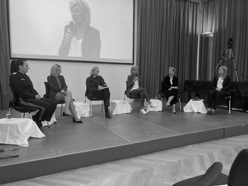 Sechs Frauen im Halbkreis sitzend auf einem Podium, eine davon ist Volksanwältin Schwarz, die ihre Rede hält.