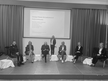 Sechs Frauen im Halbkreis sitzend auf einem Podium mit Bildschirmpräsentation im Hintergrund. Moderatorin stehend in der Mitte hinter den Teilnehmerinnen.
