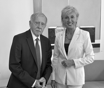 Man with grey hair and beard wearing a dark blue suit and tie, and a woman with short blond hair with an offwhite trouser suit, standing , both looking into the camera.