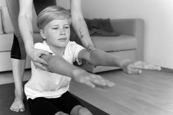 Frauenhände helfen sitzenden Jungen mit ausgestreckten Händen bei Dehnübung.